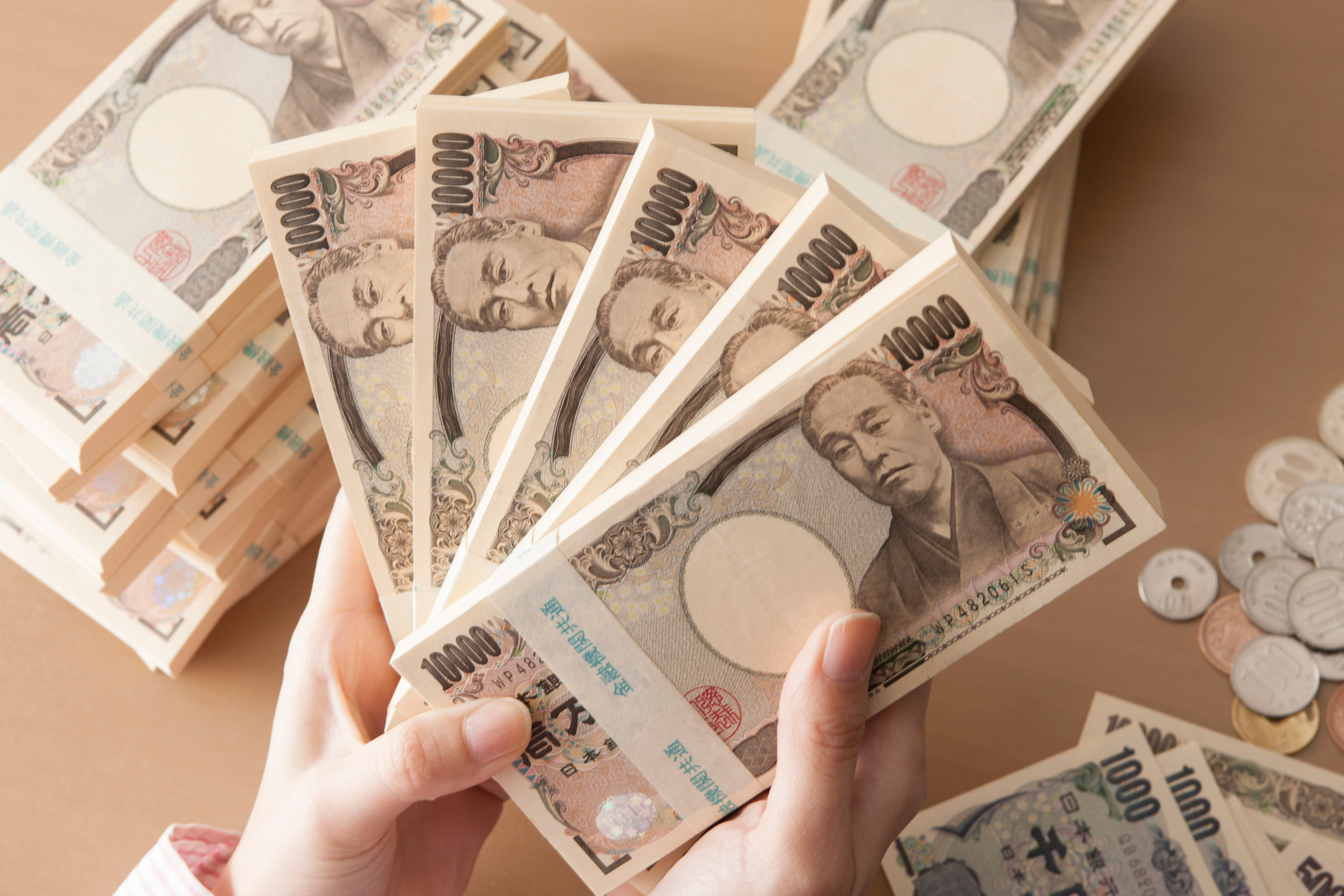 手元に数枚の1万円札が広げられ、背景には積み重ねられた日本円の札束と硬貨がある。金銭、貯蓄、豊かさのイメージ。