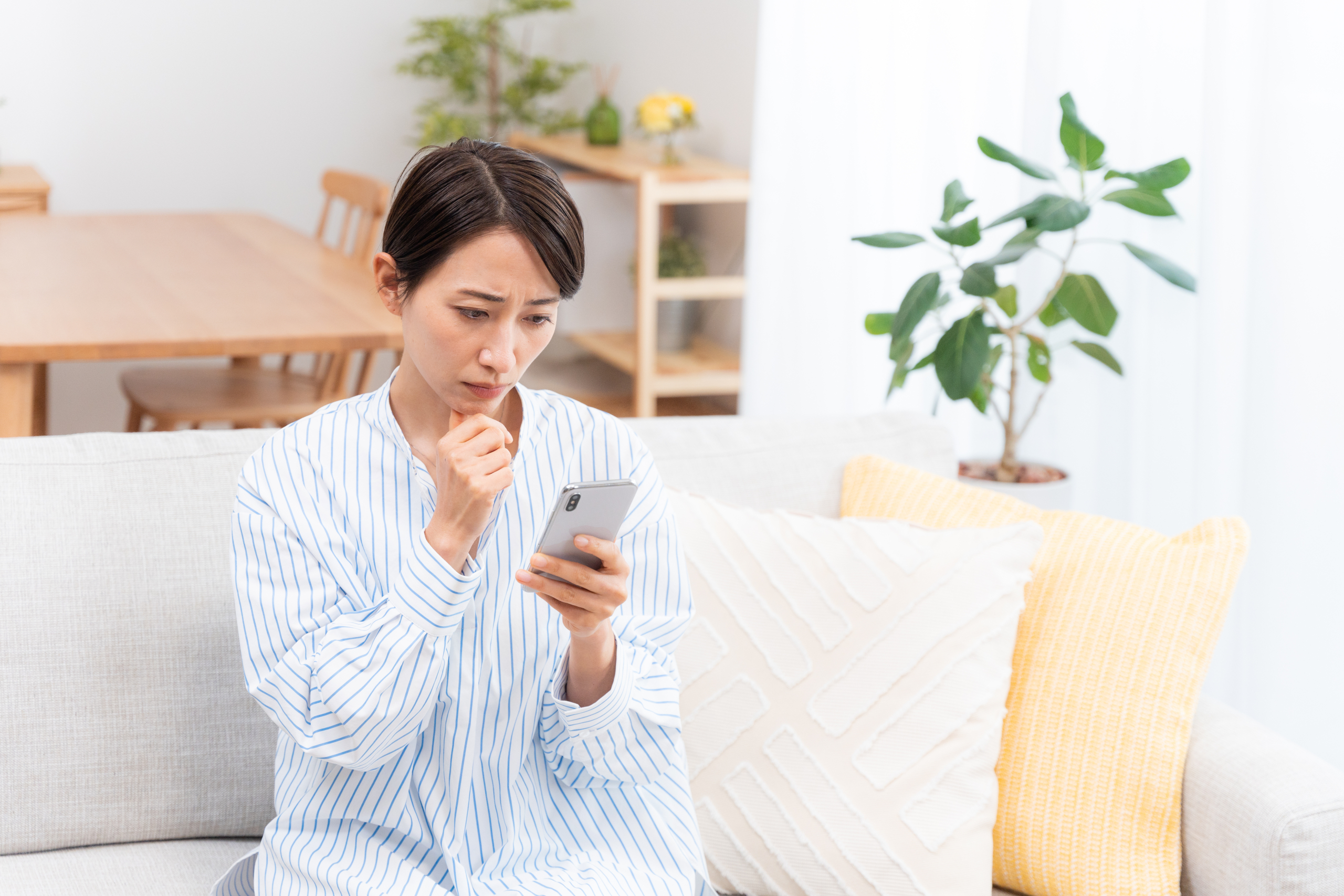リビングのソファでスマートフォンを手に、顎に手を当てて考え事をしている女性。
