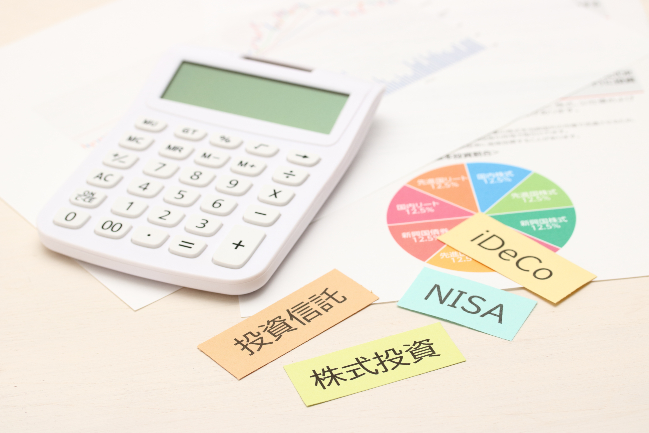 電卓とグラフ用紙の上に、投資信託、株式投資、NISA、iDeCoと書かれた付箋が置かれ、資産運用の検討を示唆する画像。