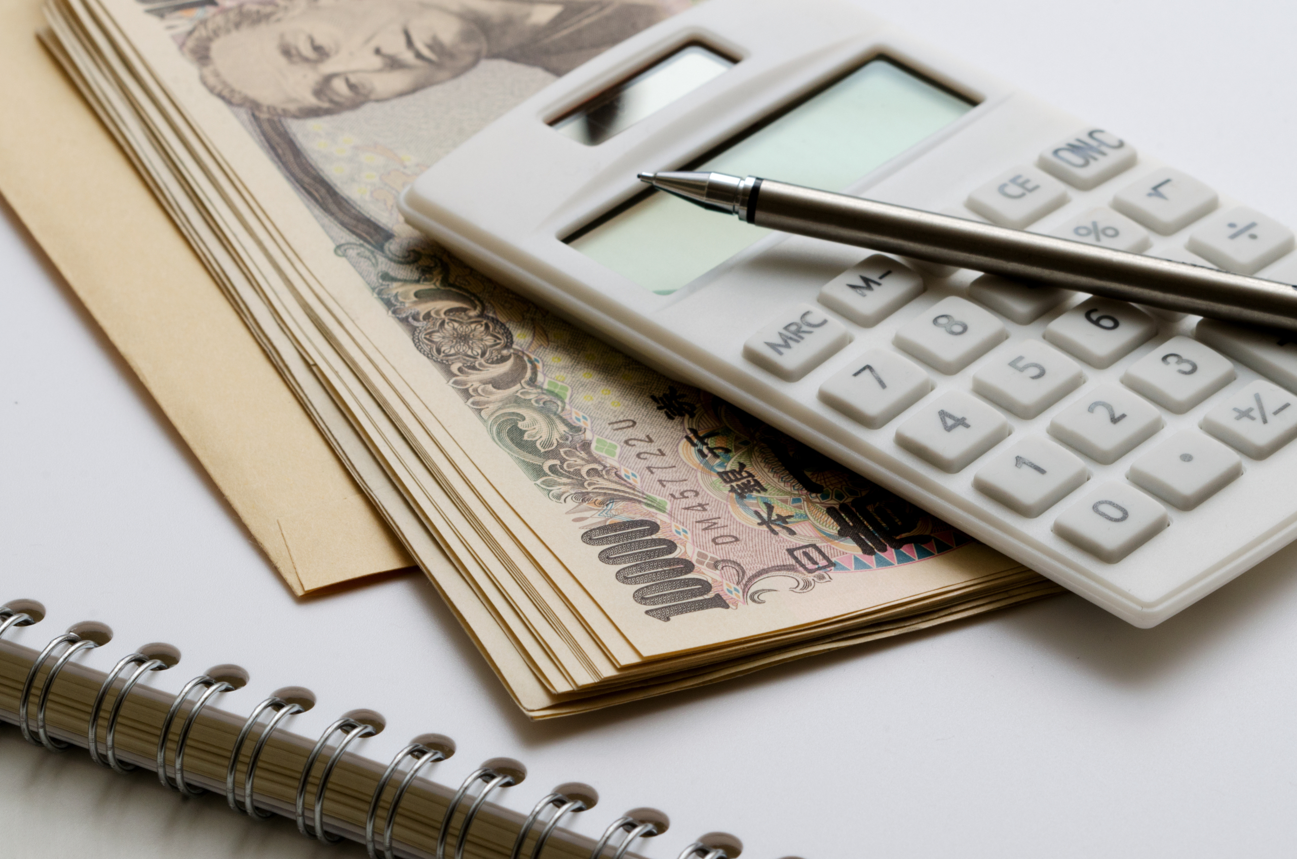 日本の1万円札の束、白い電卓、ペン、ノートが写っており、お金の計算や家計管理のイメージ。