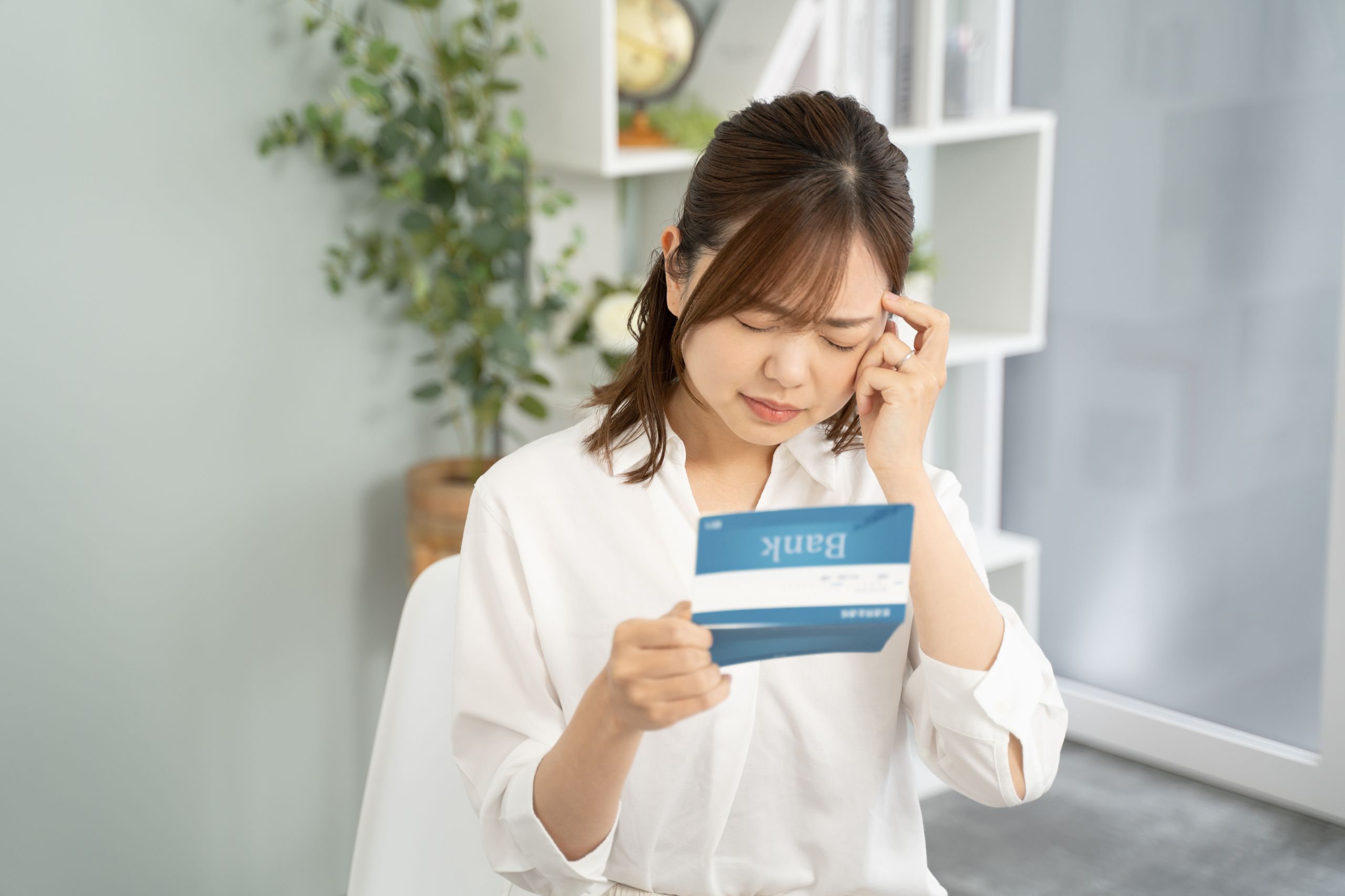 銀行の通帳（Bank）を手にし、額に手を当てて考え込んでいる女性。